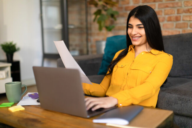 Happy spanish female real estate agent sitting at table with laptop and notepads, holding sheet of paper, writing a plan for summer real estate business.