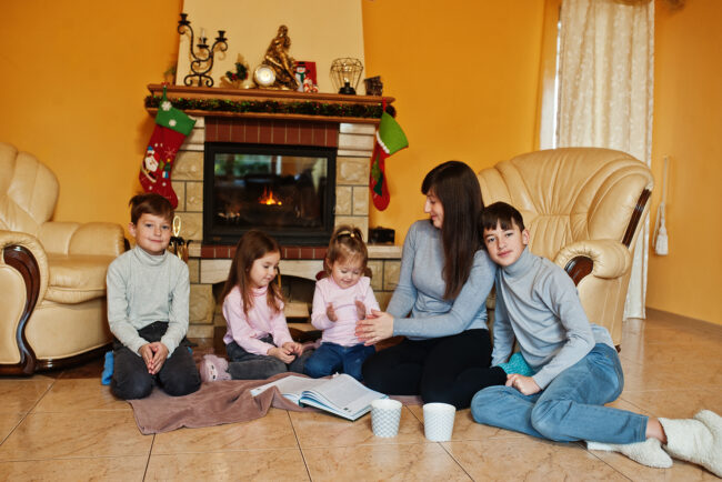 Happy young large family by a fireplace in warm living room on winter day. Mother with four kids at home.