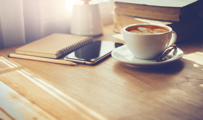 Closeup latte art coffee in white cup with smartphone and notebook on wooden table with sunlight through the window. Vintage color tone. Real estate books