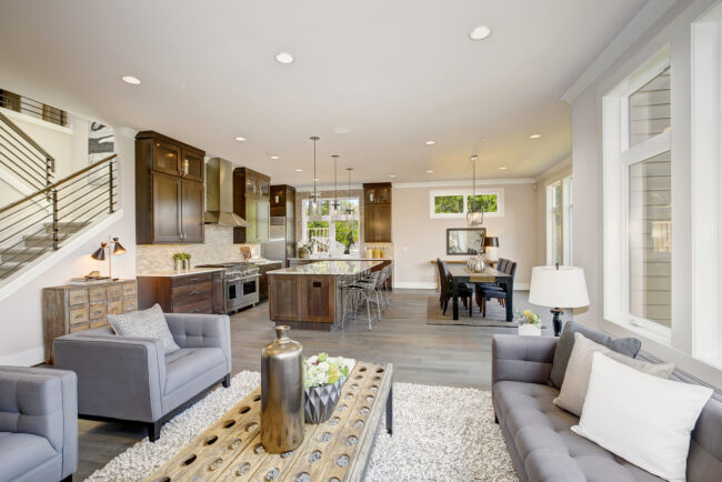 Luxurious new construction home with combination of modern style and traditional details. Kitchen features high-end appliances great room boasts gray tufted sofa and rustic wood cocktail table.